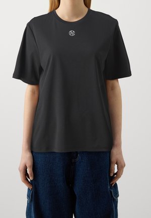 Person iført en ensfarvet sort t-shirt med et lille hvidt rundt logo midt på brystet og løstsiddende blå cowboybukser.