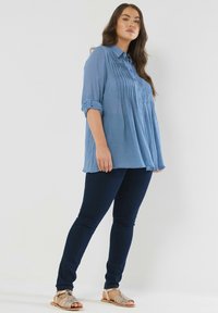 Donna in piedi con le mani rilassate, indossa una blusa plissettata blu, jeans skinny scuri e sandali aperti beige su uno sfondo semplice.