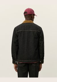 Black denim jacket with a brown corduroy collar, visible stitching, and side pockets. Worn with a maroon cap, featuring a relaxed fit.
