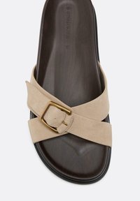 Sandale slide avec semelle intérieure marron foncé et brides croisées en suède beige avec boucle dorée sur fond blanc.