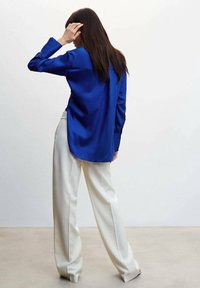 Chemise en satin bleu à manches longues, associée à un pantalon large crème, vue de dos sur fond neutre.