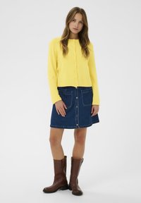 Cardigan jaune en tricot à manches longues et col rond, associé à une jupe en denim bleu avec poches avant et fermeture à boutons. Bottes hautes marron jusqu'aux genoux.