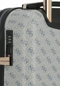 Esquina de maleta rígida con logotipo estampado, cremallera negra, cerradura combinada de tres dígitos integrada y asa de equipaje en tono dorado.