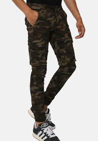 Pantalons cargo de camouflage en vert foncé et marron, comportant de multiples poches, un design slim et des poignets ajustables. Portés avec des baskets noires.