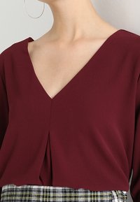 Blusa bordeaux con scollatura a V profonda e dettagli di pieghe sottili, realizzata in tessuto leggero. Abbinata a una gonna a quadri con tonalità di verde e giallo.