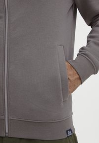 Siva zip-up dukserica s bočnim džepovima, rebrastim manžetama i porubom. Izrađena od teksturirane tkanine. Ima malu oznaku logotipa na dnu.