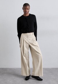 Cardigan cropped nero e morbido con chiusure a bottoni abbinato a pantaloni beige a gamba larga. Il modello indossa scarpe flat nere, in piedi su uno sfondo grigio.