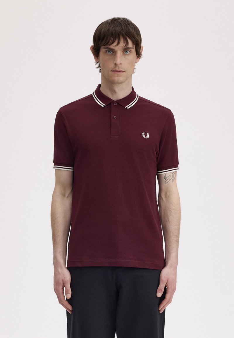 Mladić u bordo polo majici s bijelim obrubom i crnim hlačama, stoji pred jednostavnom svijetlom pozadinom.