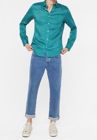 Modrý tričko s dlouhým rukávem, zapínáním na knoflíky a měkkou texturou, doplněné modrými riflemi s manžetami a béžovými teniskami, vytvářející neformální outfit.