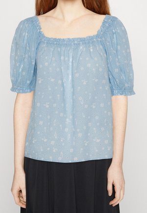 Femme portant un chemisier à manches bouffantes bleu clair à motifs floraux avec un col carré élastique et une jupe noire, les bras détendus le long du corps.