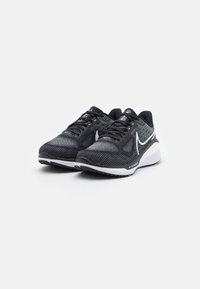 Baskets de sport noires avec un upper en mesh texturé, des accents blancs et une semelle en caoutchouc robuste. Présente des lacets et une pointe arrondie.