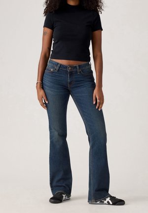 Kvinde iført mørkeblå bootcut-jeans, sort cropped rullemelontrøje og sorte sneakers med hvide striber, stående mod en ensfarvet baggrund.