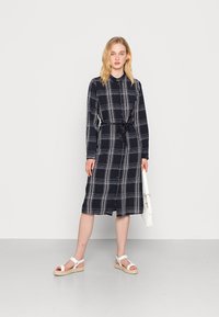 Vestido xadrez azul-marinho com cintura cintada, frente de botões e mangas longas. Modelo usa sandálias brancas e segura uma bolsa branca.