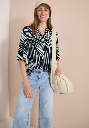 Femme souriante portant une chemise à imprimé zèbre noir et blanc, un jean bleu clair, une casquette beige, tenant un sac à bandoulière matelassé beige contre un mur beige.