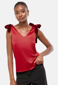 Piros selyem ottónyú, V-kivágással, mely vállain bársonyos masnikkal díszített, sima, fényes textúrával. Fekete nadrágok viselve alatta.