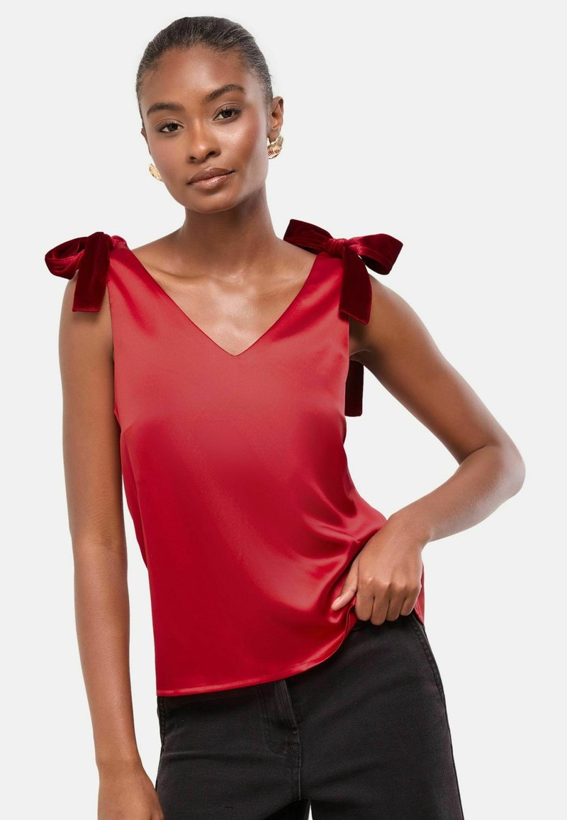 Piros selyem ottónyú, V-kivágással, mely vállain bársonyos masnikkal díszített, sima, fényes textúrával. Fekete nadrágok viselve alatta.