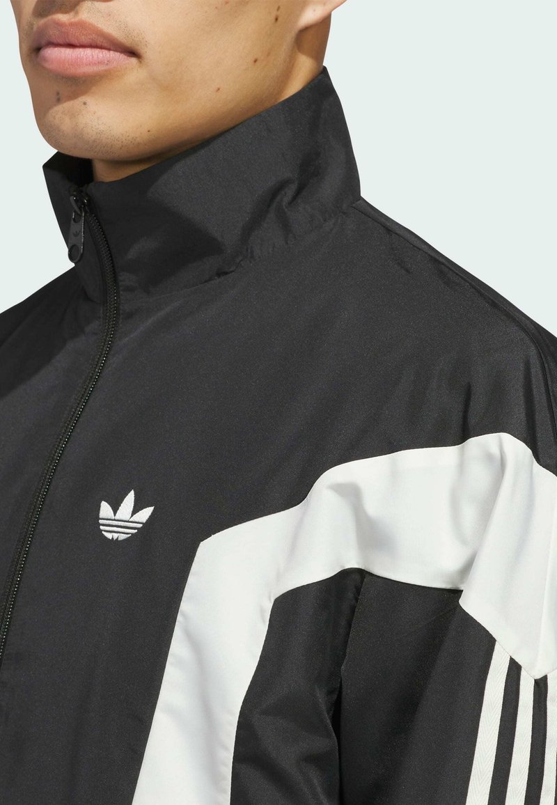 Schwarze Zip-Jacke mit hohem Kragen, mit weißen Einsätzen und dem Adidas-Logo auf der linken Seite; aus glattem, leichtem Material gefertigt.