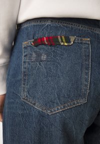 Jeans i denim med en mörkblå tvätt, med en bakficka prydd med en vävd, flerfärgad rutig detalj och synliga sömdetaljer.