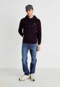 Felpa bordeaux con cordini e tasca frontale; abbinata a jeans azzurri chiari e sneakers beige. Cappellino grigio con stampa sulla parte anteriore.