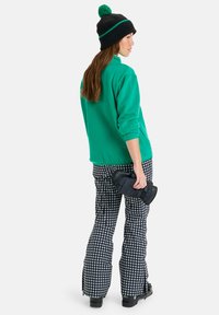 Casaco de fleece verde, gorro de malha preto com pompom verde, calças pretas e brancas estampadas, segurando um objeto acolchoado preto.