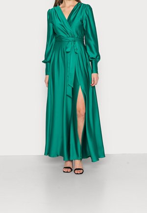Vrouw draagt een lange teal satijnen wikkeljurk met poflange mouwen, een V-hals, een taillekoord en een hoge frontsplit, gecombineerd met zwarte open-toe hakken.