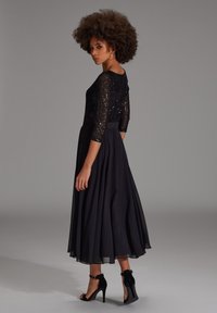 Robe noire avec un haut en sequins et des manches trois-quarts, jupe ample en forme de A, sandales à bride cheville et texture douce.