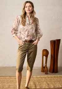 Nuori nainen pitkissä hiuksissa, yllään vaalea kukkakuosinen pusero ja khakinväriset vyölliset kapenevat housut, seisomassa sisätiloissa puisien veistosten lähellä matolla.