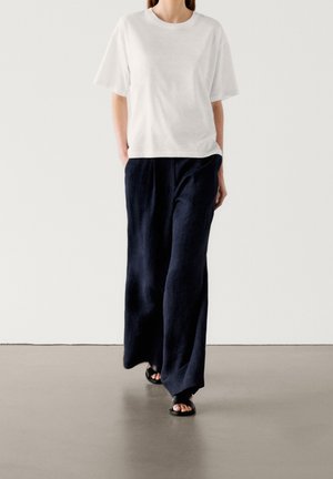 Person trägt ein weites weißes kurzärmeliges Hemd, weite schwarze Hosen und schwarze Sandalen mit offenen Zehen und geht auf einem glatten Boden.