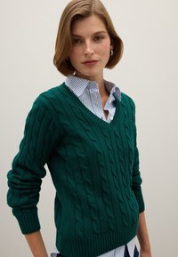Tealfarbener Kabelstrickpullover mit V-Ausschnitt, gerippten Bündchen und Saum, über einem hellblauen gestreiften Hemd mit Kragen getragen. Glatte Textur.
