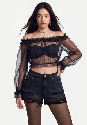 Femme portant un haut noir transparent épaules dénudées avec un bandeau noir, un short en jean noir déchiré et des boucles d'oreilles et un collier en forme d'étoile.