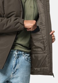 Brun polstret jakke med lynlås, der har en glat overflade og indvendige lommer. Bæres over en grøn skjorte og lyseblå jeans.