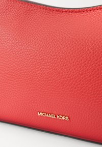 Sac à main en cuir rouge avec une finition texturée, arborant une plaque logo en or. Le design est épuré avec un bord supérieur arrondi.