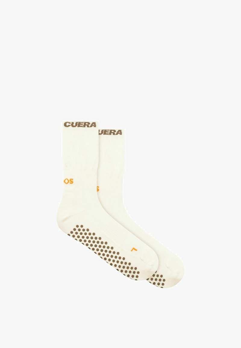 Paire de chaussettes blanches montantes avec des points d'adhérence marron sous les semelles et un texte marron près du bord supérieur.