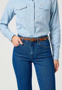 Camisa de cuadros en azul claro con dos bolsillos en el pecho, combinada con vaqueros azul oscuro y un cinturón de cuero marrón con una hebilla de plata.