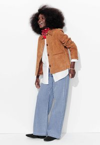 Veste en daim marron clair avec de grands boutons marron sur une chemise blanche, associée à un pantalon large rayé bleu et blanc et des chaussures noires cloutées. Foulard rouge en accent.