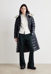 Langer schwarzer Puffer-Mantel mit hohem Kragen, hellgrauem Innenfutter, kombiniert mit einem beigen Hemd mit Knöpfen und dunkelgrünen, weitem Hosen. Schwarze Plateauschuhe.