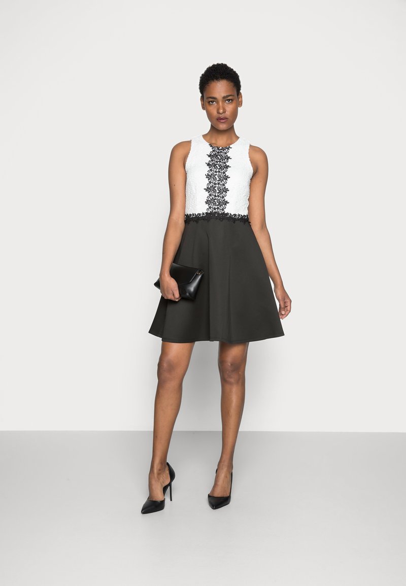 Schwarz-weißes Kleid mit einem strukturierten Oberteil mit Spitzenbesatz, einem ausgestellten schwarzen Rock, kombiniert mit schwarzen hohen Absätzen und einer Clutch.