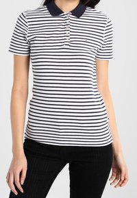 Polo tričko s pruhy v námořnické modré a bílé barvě, s námořnickým límečkem, krátkými rukávy a pěticí knoflíků. Obsahuje malé logo na hrudi.