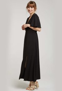 Robe maxi noire avec des manches courtes évasées, décolleté en V et fente à l'avant. Tissu doux avec une taille ajustée, associée à des sandales à talons carrés beiges.