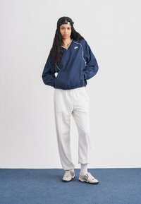 Tengerészkék széldzseki rugalmas mandzsettákkal, fehér szegéllyel és Nike logóval; világosszürke, rugalmas derékpántos nadrág; bézs sportcipők fekete részletekkel.