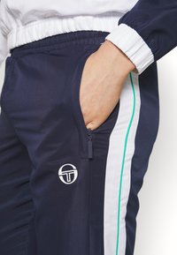 Navy track pants with white side stripes and teal accents. Features a zippered pocket and a circular logo on the left leg.