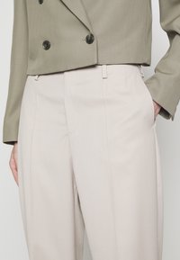 Pantalon beige clair sur mesure avec plis avant, associé à un blazer croisé olive raccourci avec des boutons noirs.