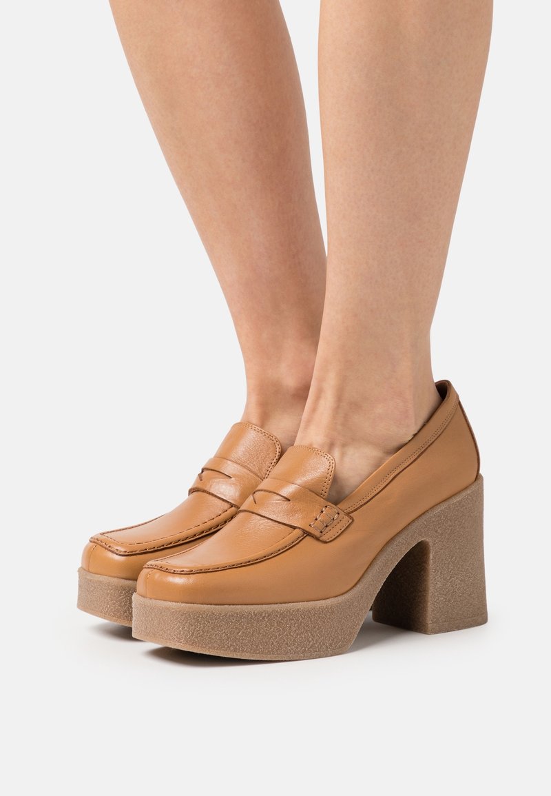 Mocassins à plateforme en cuir tan avec des talons larges, présentant des détails cousus et une finition lisse. Les semelles sont texturées et épaisses.