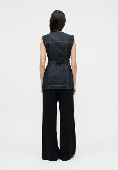 Γυναίκα με μακριά σκούρα μαλλιά, φορούν αμάνικο σκούρο τζιν γιλέκο με ζώνη και φαρδιά μαύρη παντελόνα, στέκεται με την πλάτη γυρισμένη πάνω σε λευκό φόντο.