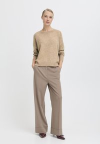 Beżowy sweter w sploty z okrągłym dekoltem i długimi rękawami, w zestawieniu z jasnobrązowymi spodniami szerokimi w nogawkach z mankietem oraz ciemnymi butami.
