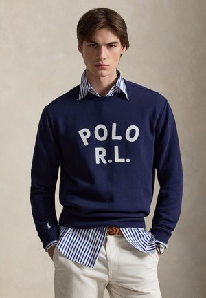 Noormees kannab tumesinist „POLO R.L.” pusa sinise-valge triibulise särgi ja valgete pükstega ning pruun vöö, käed taskutes.