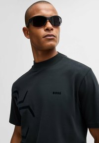 T-shirt noir à col rond avec une texture lisse, présentant un logo et un motif abstrait. Accentué par de lunettes de soleil sombres élégantes.