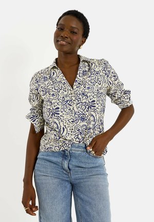 Mujer sonriente con el pelo corto, que lleva una camisa blanca y azul con botones y vaqueros azul claro, posando con una mano en el bolsillo.