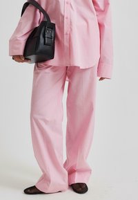 Roza oversized srajca in usklajene hlače iz lahkotnega materiala, v kombinaciji s trdno črno torbico in mrežastimi črnimi čevlji.