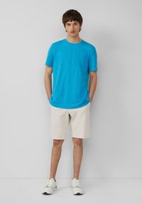 Jasnoniebieska koszulka z krótkim rękawem o teksturowanym wykończeniu, w zestawieniu z kremowymi szortami i białymi butami sportowymi. Prosty, codzienny strój.